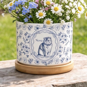 Puede incluir: Una maceta cilíndrica de cerámica blanca con un patrón floral azul y un marco ovalado central que contiene la ilustración de un gato y el nombre Nova. La maceta descansa sobre un plato de bambú y está llena de flores silvestres blancas y azules.
