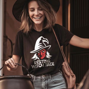 May include: A black short-sleeved t-shirt featuring a graphic of a red-faced witch wearing a pointed hat, with the white text "RESTING WITCH FACE" printed underneath. The design has a distressed, weathered appearance on the fabric.