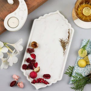 May include: A white marble tray with a scalloped edge, decorated with scattered dried red flower petals, berries, and a small brown twig. The tray is positioned among white plumeria flowers, a glass of yellow liquid with a lemon slice, and a patterned cloth.