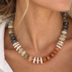 May include: A beaded necklace featuring a gradient of smooth, round, disc-shaped beads in shades of grey, cream, peach, and orange, separated by small gold-toned spacers. The necklace is worn around the neck, complemented by small gold hoop earrings.