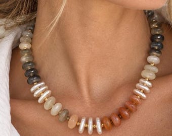 May include: A beaded necklace featuring a gradient of smooth, round, disc-shaped beads in shades of gray, cream, peach, and orange, separated by small gold-toned spacers. The necklace is worn around the neck, complemented by small gold hoop earrings.