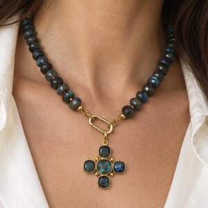 Pu&ograve; includere: Una collana con una fila di perline di labradorite scure e iridescenti e una chiusura in metallo dorato. Il ciondolo ha un design a croce composto da cinque pietre di labradorite quadrate incastonate in metallo dorato, appeso alla chiusura.