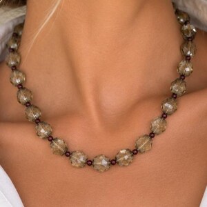 May include: A beaded necklace featuring faceted, translucent grey-brown beads alternating with small, dark red spherical beads. The necklace is worn around the neck, resting against a white collared shirt, and is paired with small gold hoop earrings.