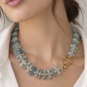 Peut inclure: Un collier compos&eacute; d'une alternance de perles rectangulaires en pierre bleu clair et de petites perles sph&eacute;riques blanches. Le bijou est ferm&eacute; par un fermoir &agrave; bascule en m&eacute;tal dor&eacute;, form&eacute; d'un cadre carr&eacute; et d'une barre en T &agrave; l'avant.