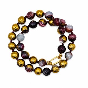 May include: A beaded necklace featuring alternating faceted metallic gold beads and smooth, round, multi-colored agate beads in shades of deep purple, maroon, and grey. The necklace is secured with a polished gold-tone oval link clasp.
