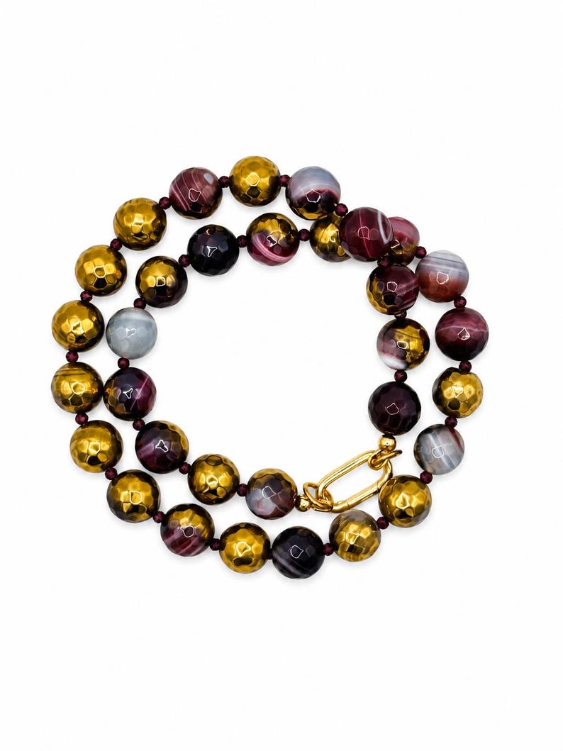 May include: A beaded necklace featuring alternating faceted metallic gold beads and smooth, round, multi-colored agate beads in shades of deep purple, maroon, and grey. The necklace is secured with a polished gold-tone oval link clasp.