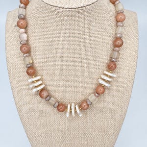 May include: A beaded necklace featuring round peach-coloured stones, translucent square beads, and small grey spacer beads. The design includes clusters of elongated white stick pearls accented with small gold-toned metal beads throughout the strand.