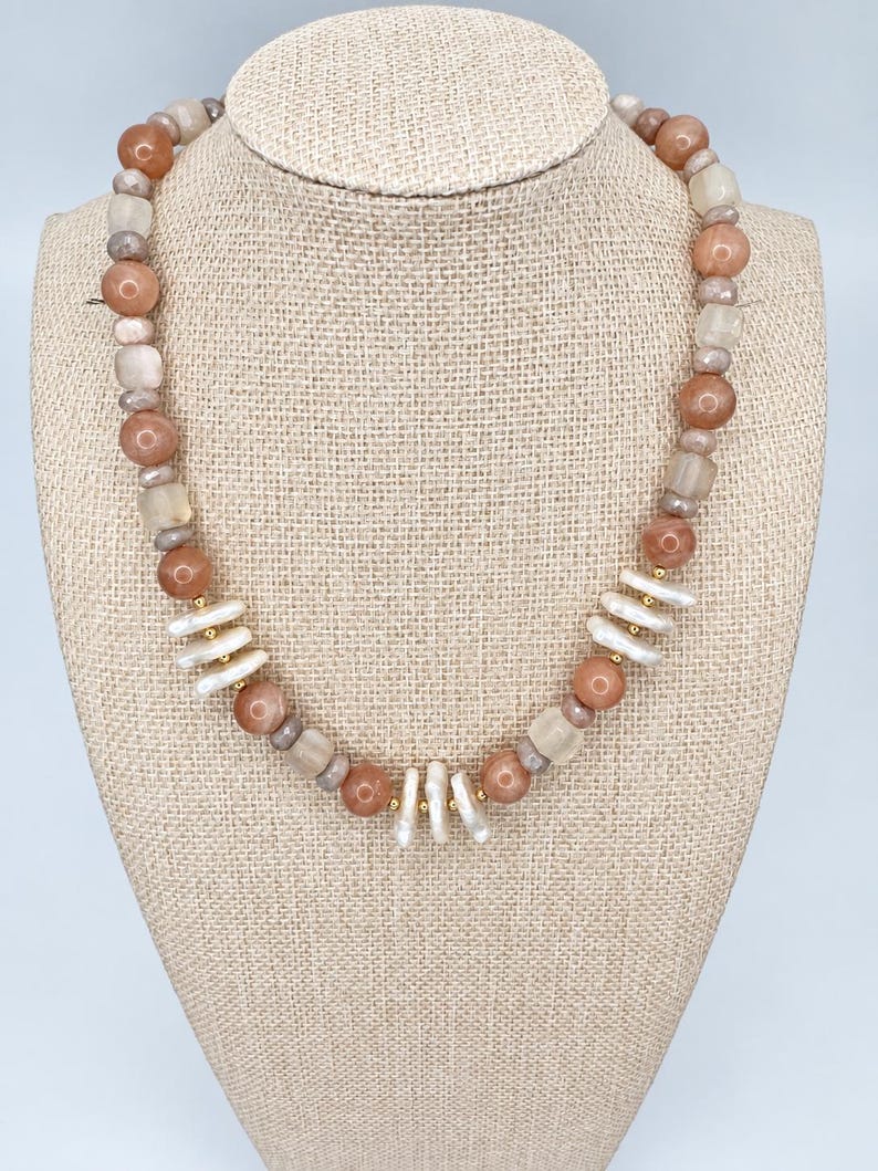 May include: A beaded necklace featuring round peach-coloured stones, translucent square beads, and small grey spacer beads. The design includes clusters of elongated white stick pearls accented with small gold-toned metal beads throughout the strand.