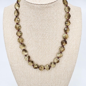 May include: A necklace featuring faceted, translucent, brownish-grey beads arranged in a repeating pattern with small, round, deep red spacer beads between each larger bead. The necklace is displayed in a circular shape on a neutral-colored stand.