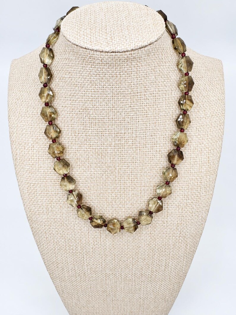 May include: A necklace featuring faceted, translucent, brownish-grey beads arranged in a repeating pattern with small, round, deep red spacer beads between each larger bead. The necklace is displayed in a circular shape on a neutral-colored stand.