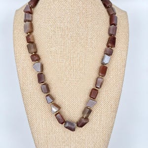 May include: A necklace featuring a series of irregular, tumbled stone beads in shades of brown, tan, and gray. The beads are separated by small, round gold-toned metal spacers, creating a continuous strand of earthy, polished natural stones.
