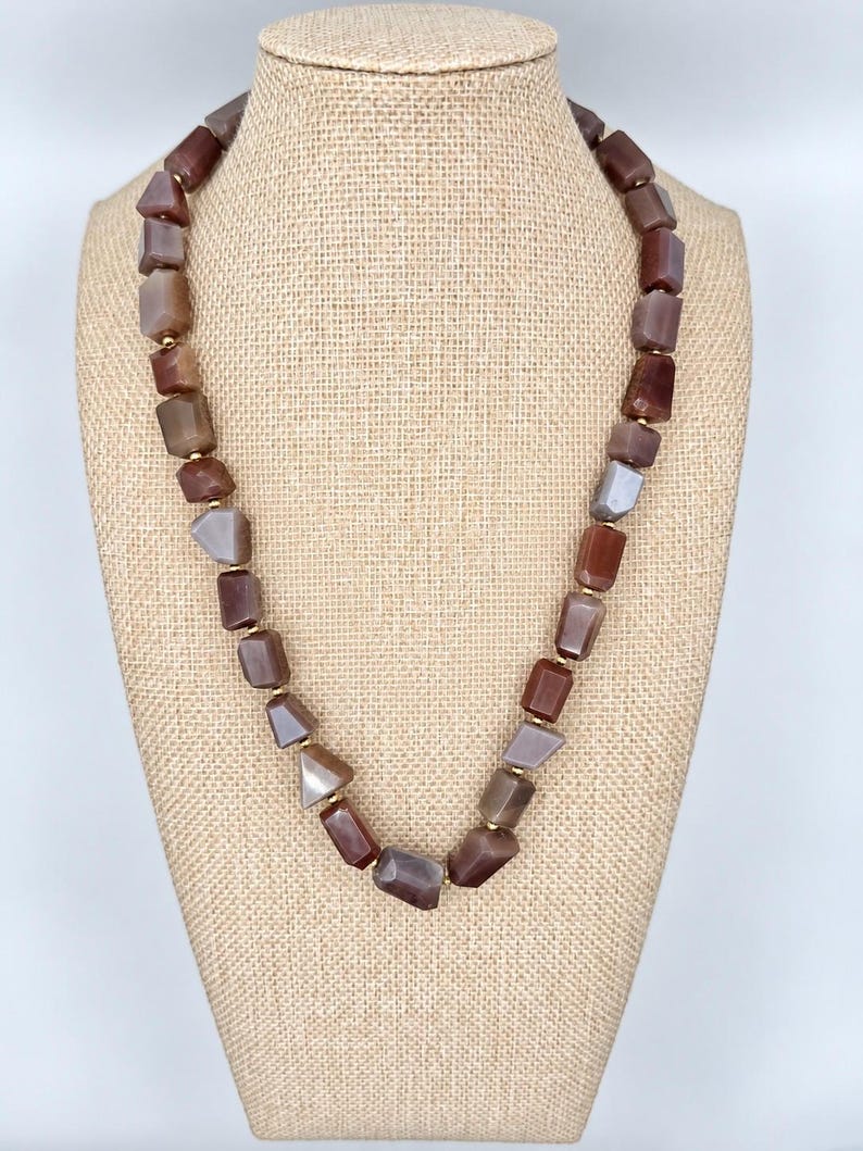 May include: A necklace featuring a series of irregular, tumbled stone beads in shades of brown, tan, and gray. The beads are separated by small, round gold-toned metal spacers, creating a continuous strand of earthy, polished natural stones.