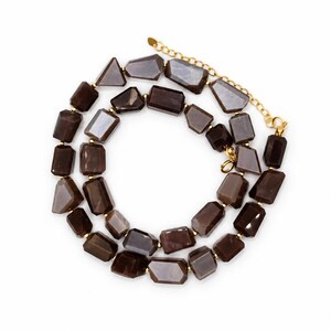 May include: A necklace featuring two strands of faceted, dark brown, rectangular-shaped beads. The beads are separated by small gold-toned metal spacers. The necklace includes a gold-toned metal chain extender and a lobster claw clasp.