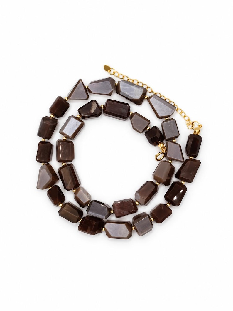 May include: A necklace featuring two strands of faceted, dark brown, rectangular-shaped beads. The beads are separated by small gold-toned metal spacers. The necklace includes a gold-toned metal chain extender and a lobster claw clasp.