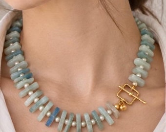 May include: A necklace featuring rectangular, light blue stone beads alternating with small, round white pearls. The piece is secured with a gold-toned metal toggle clasp, which consists of a square frame and a T-bar, positioned at the front.