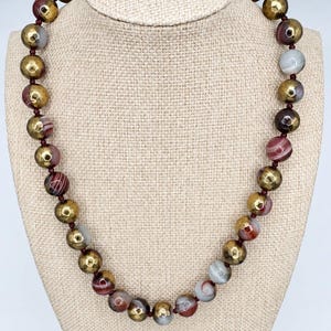 May include: A beaded necklace featuring alternating round beads in a metallic gold finish with a faceted texture and smooth, multi-colored banded agate stones in shades of brown, red, white, and gray, all strung on a dark red cord.