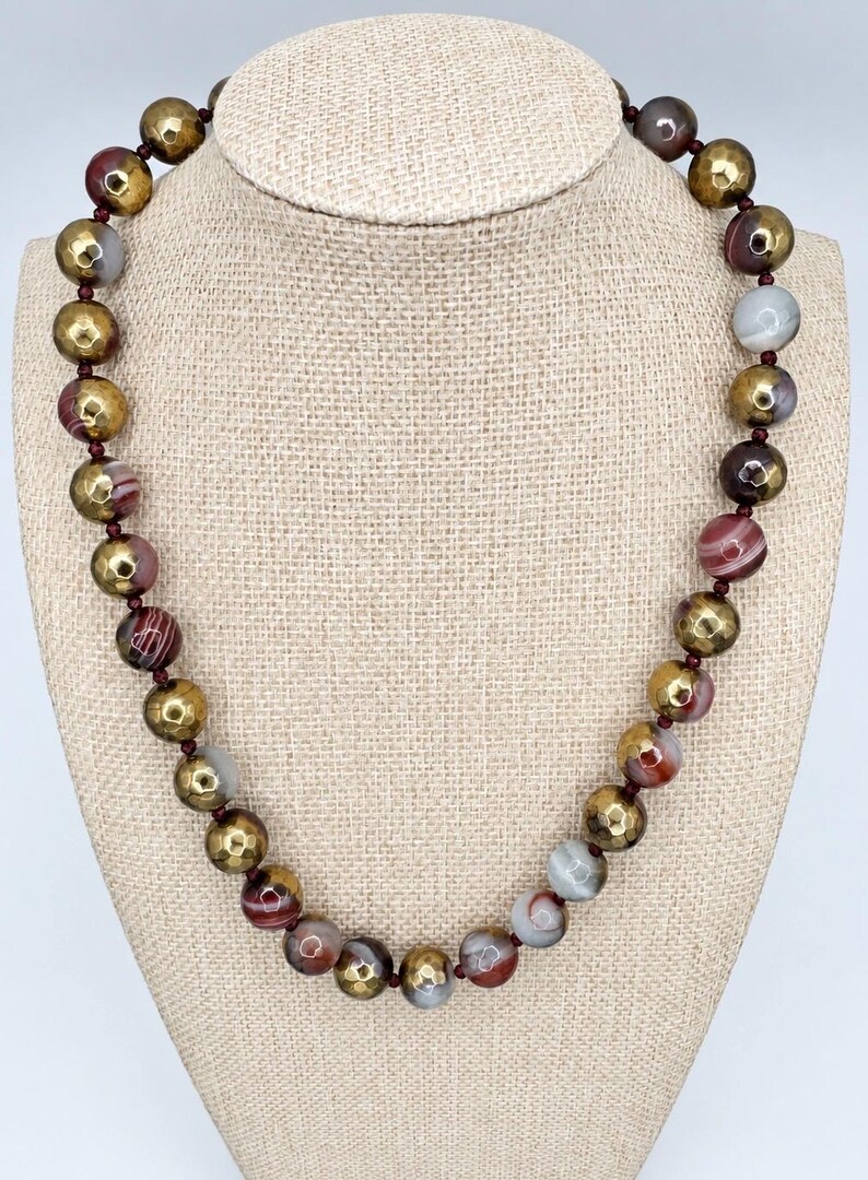 May include: A beaded necklace featuring alternating round beads in a metallic gold finish with a faceted texture and smooth, multi-colored banded agate stones in shades of brown, red, white, and gray, all strung on a dark red cord.