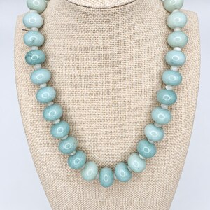 May include: A single-strand necklace featuring smooth, round, pale seafoam green beads. The beads are arranged in a uniform sequence, separated by small, translucent white spacer beads, creating a consistent circular pattern around the neck.