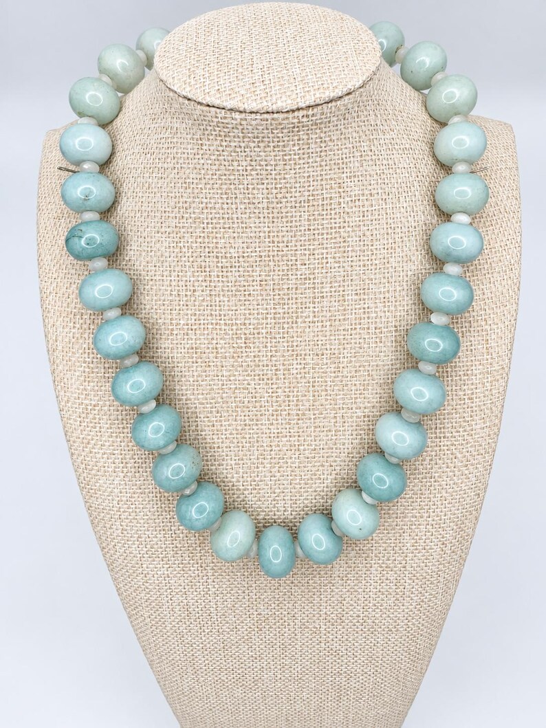 May include: A single-strand necklace featuring smooth, round, pale seafoam green beads. The beads are arranged in a uniform sequence, separated by small, translucent white spacer beads, creating a consistent circular pattern around the neck.
