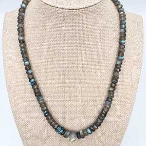 May include: A necklace composed of numerous small, faceted labradorite beads in shades of grey and brown, featuring iridescent blue and gold flashes. The beads are strung in a continuous circular pattern, with a slightly larger, central faceted bead.