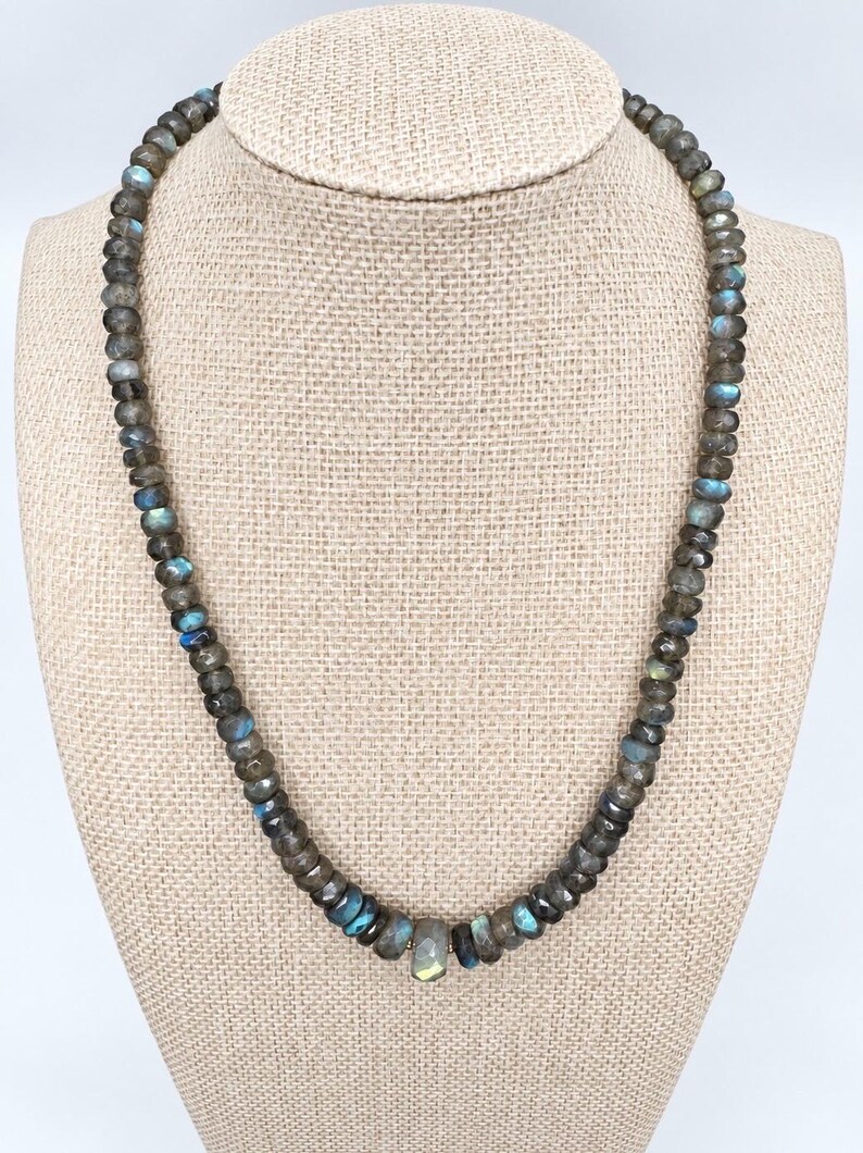 May include: A necklace composed of numerous small, faceted labradorite beads in shades of grey and brown, featuring iridescent blue and gold flashes. The beads are strung in a continuous circular pattern, with a slightly larger, central faceted bead.