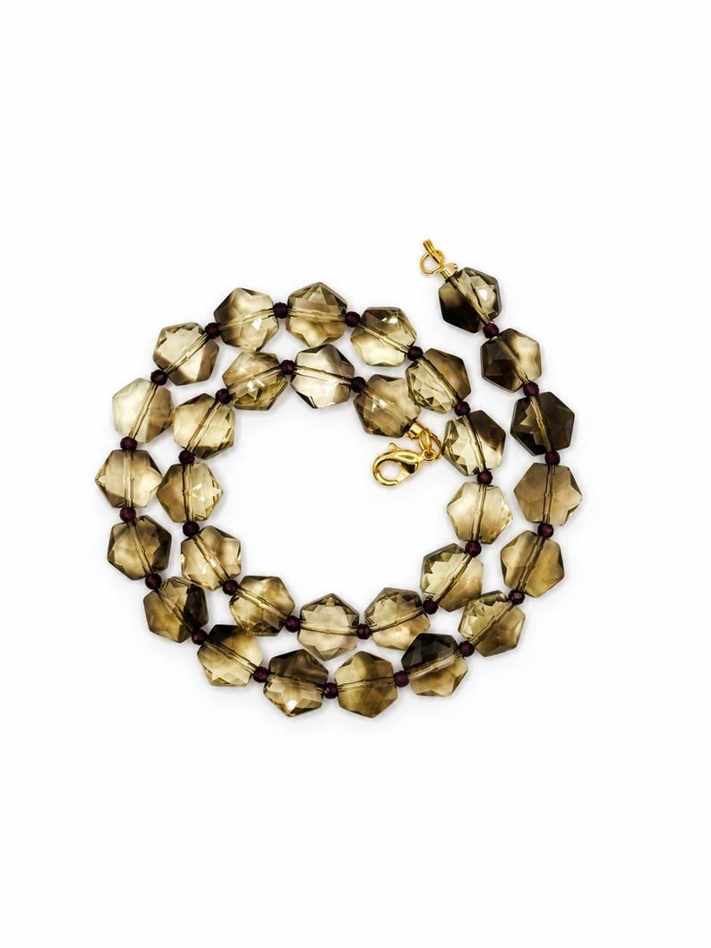 May include: A necklace featuring faceted, hexagonal smoky quartz beads in a translucent brown hue, interspersed with small, dark maroon spherical spacer beads. The strand is secured with a gold-toned metal lobster claw clasp and jump ring.