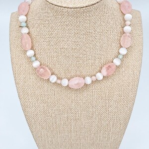 May include: A beaded necklace featuring alternating light pink faceted oval beads and smooth white round beads. The design includes small, pale green accent beads interspersed throughout the strand, creating a balanced and symmetrical pattern of soft colors.