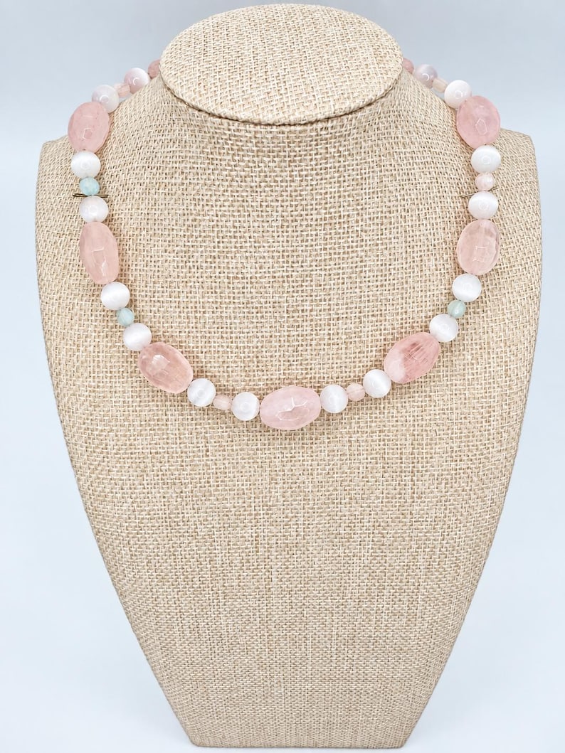 May include: A beaded necklace featuring alternating light pink faceted oval beads and smooth white round beads. The design includes small, pale green accent beads interspersed throughout the strand, creating a balanced and symmetrical pattern of soft colors.