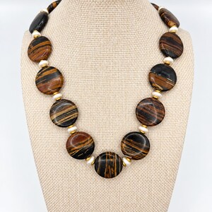 The Sutton necklace displayed on a beige fabric jewelry bust against a pale background, with round brown-and-black striped beads, creamy pearls and gold-tone spacers.