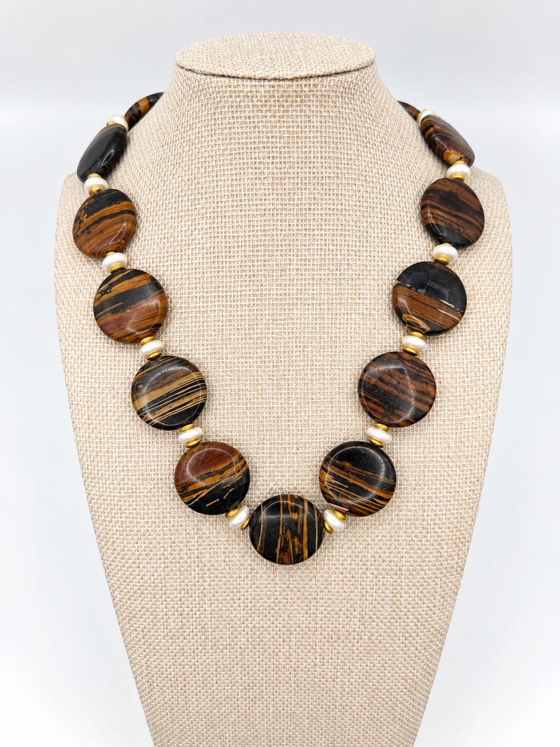 The Sutton necklace displayed on a beige fabric jewelry bust against a pale background, with round brown-and-black striped beads, creamy pearls and gold-tone spacers.