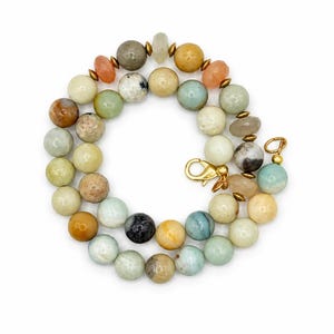 May include: A beaded necklace featuring a variety of round, smooth, and faceted stones in shades of beige, tan, light blue, green, and grey. The beads are interspersed with small metallic gold-toned spacers and finished with a gold-toned lobster clasp.
