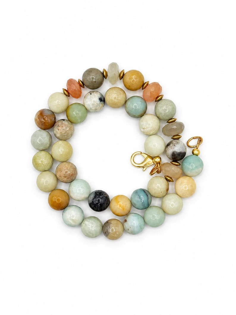 May include: A beaded necklace featuring a variety of round, smooth, and faceted stones in shades of beige, tan, light blue, green, and grey. The beads are interspersed with small metallic gold-toned spacers and finished with a gold-toned lobster clasp.