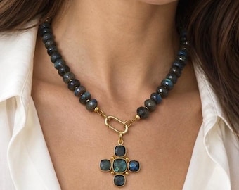 May include: A necklace featuring dark grey labradorite beads with iridescent blue flashes, connected by a gold-toned oval clasp to a cross-shaped pendant. The pendant consists of five square-cut labradorite stones set in gold-toned metal frames.