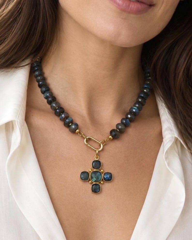 The Monaco necklace worn with an open white shirt: dark labradorite beads, a gold-colored front ring and a cross-shaped blue-green stone pendant.