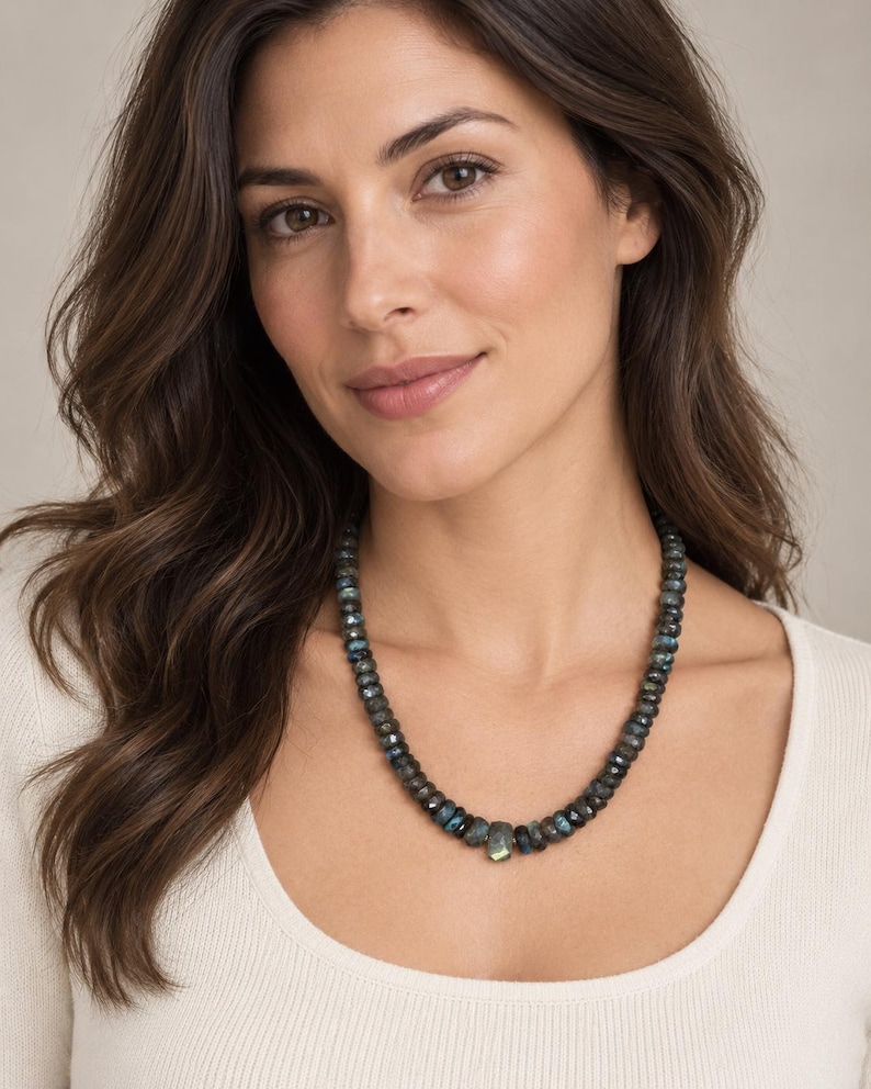 May include: A necklace composed of faceted, dark grey labradorite beads with iridescent blue and green flashes. The beads are round and uniform in size, except for a single, slightly larger, rectangular-shaped bead positioned at the centre.