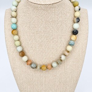 May include: A beaded necklace featuring a variety of round, multi-colored gemstone beads in shades of cream, tan, light blue, grey, and orange. The beads are interspersed with small, thin, metallic gold-toned spacer discs throughout the strand.
