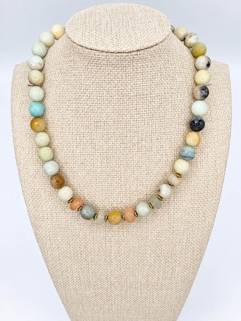 May include: A beaded necklace featuring a variety of round, multi-colored gemstone beads in shades of cream, tan, light blue, grey, and orange. The beads are interspersed with small, thin, metallic gold-toned spacer discs throughout the strand.