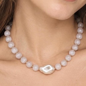 May include: A necklace featuring a series of smooth, pale pink spherical beads interspersed with small gold-toned spacers, centered with a single, large, irregular-shaped white pearl-like bead that has a lustrous, reflective surface finish.