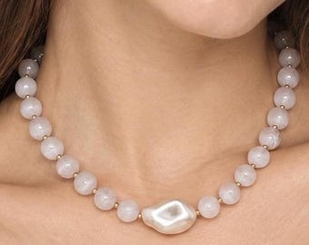 Peut inclure: Un collier composé d'une série de perles sphériques rose pâle lisses, séparées par de petites entretoises dorées, avec en son centre une unique grosse perle blanche de forme irrégulière dotée d'une finition de surface lustrée et réfléchissante.