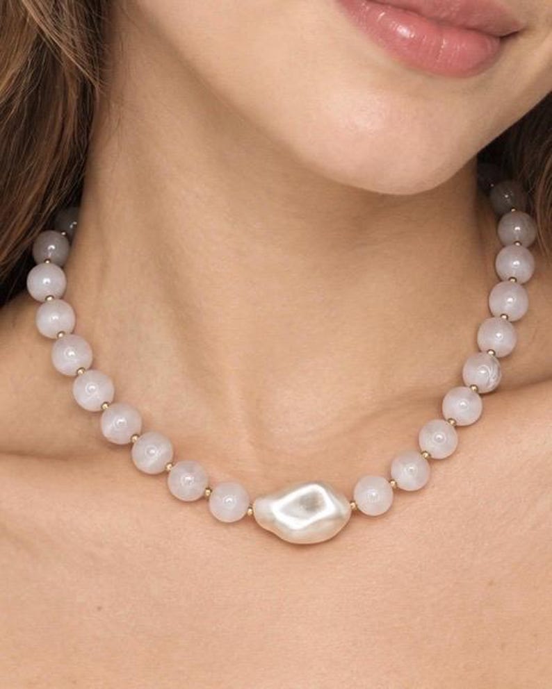 May include: A necklace featuring a series of smooth, pale pink spherical beads interspersed with small gold-toned spacers, centered with a single, large, irregular-shaped white pearl-like bead that has a lustrous, reflective surface finish.