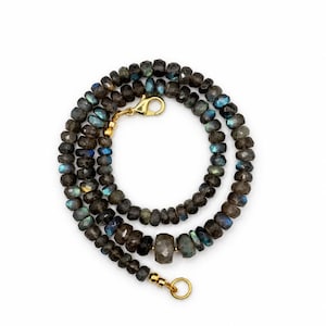 May include: A necklace composed of faceted, dark grey labradorite beads that exhibit iridescent blue and gold flashes. The strand features a gold-toned metal lobster claw clasp and a circular ring connector, with a larger central bead.