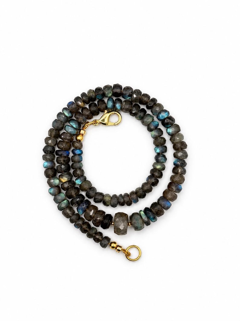 May include: A necklace composed of faceted, dark grey labradorite beads that exhibit iridescent blue and gold flashes. The strand features a gold-toned metal lobster claw clasp and a circular ring connector, with a larger central bead.