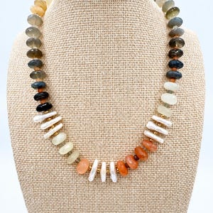 May include: A beaded necklace featuring a variety of faceted round stones in shades of grey, blue, black, and orange, interspersed with white elongated freshwater pearls and small gold-toned metal spacer beads arranged in a circular pattern.