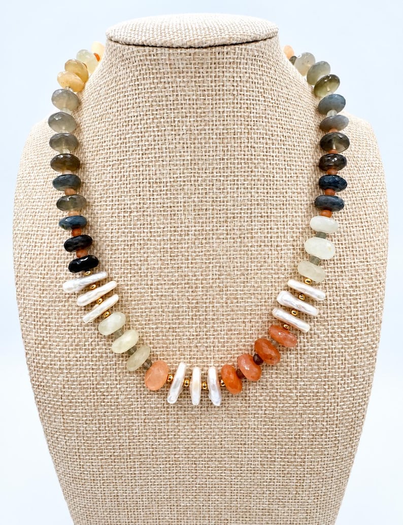 May include: A beaded necklace featuring a variety of faceted round stones in shades of grey, blue, black, and orange, interspersed with white elongated freshwater pearls and small gold-toned metal spacer beads arranged in a circular pattern.
