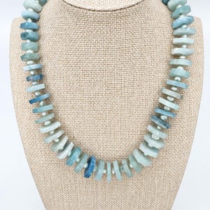 Peut inclure: Un collier compos&eacute; d'une s&eacute;rie de perles de pierre plates et irr&eacute;guli&egrave;res, dans des tons bleu clair et sarcelle, altern&eacute;es avec de petites perles d'espacement rondes blanc cass&eacute;. Les pierres varient en teinte et en opacit&eacute;.
