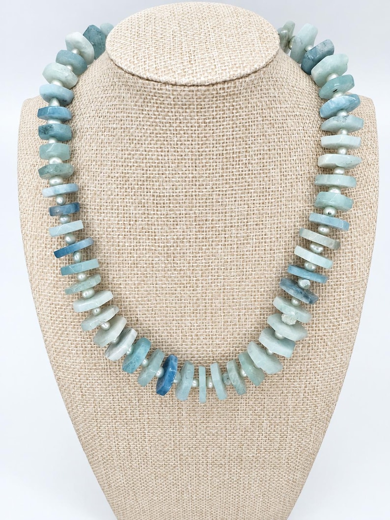 Peut inclure: Un collier compos&eacute; d'une s&eacute;rie de perles de pierre plates et irr&eacute;guli&egrave;res, dans des tons bleu clair et sarcelle, altern&eacute;es avec de petites perles d'espacement rondes blanc cass&eacute;. Les pierres varient en teinte et en opacit&eacute;.