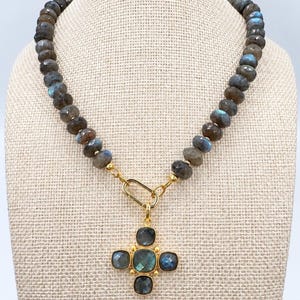 Pu&ograve; includere: Una collana con perle di labradorite rotonde e sfaccettate con riflessi blu iridescenti. Il filo &egrave; collegato a un anello ovale dorato che sostiene un ciondolo a forma di croce composto da cinque pietre di labradorite quadrate e sfaccettate.