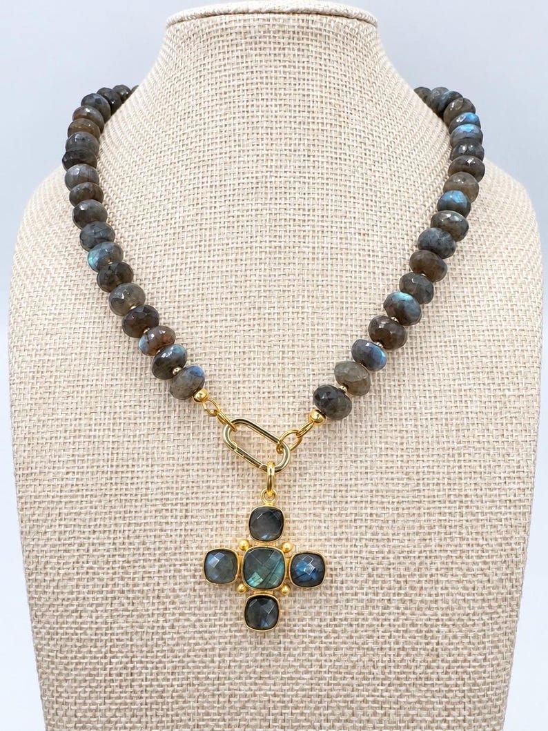 Pu&ograve; includere: Una collana con perle di labradorite rotonde e sfaccettate con riflessi blu iridescenti. Il filo &egrave; collegato a un anello ovale dorato che sostiene un ciondolo a forma di croce composto da cinque pietre di labradorite quadrate e sfaccettate.