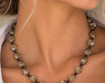 May include: A necklace featuring a series of faceted, translucent smoky gray beads alternating with small, round, dark maroon spacer beads. The beads are strung together in a single strand, creating a uniform pattern around the neck area.