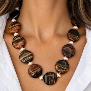 The Sutton necklace, with large round brown-and-black striped beads, small creamy white pearls and gold-tone accents, worn with a white open-collar shirt.